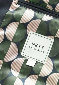 Tissu avec des motifs circulaires à demi-rayures vertes et beiges, et une étiquette carrée blanche cousue portant l'inscription "NEXT TAILORING".