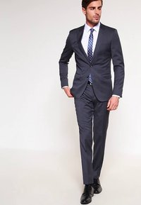 Costume marine à coupe ajustée, fermeture à un bouton, revers crantés, associé à une chemise blanche et une cravate à motifs. Chaussures habillées noires.