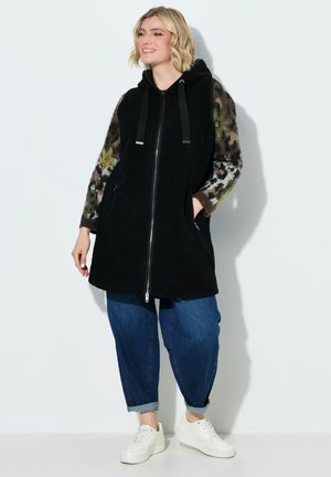 Kvinde iført sort vest med lynlås og hætte over camouflagefarvede ærmer, blå opkørte jeans og hvide sneakers, stående op ad en hvid væg.