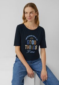 Femme aux cheveux blonds portant une chemise marine avec le texte "Je suis tellement excitée pour toutes les bonnes choses à venir", assise contre un fond gris uni.