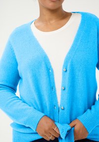 Helles blauer Cardigan aus flauschigem Stoff, mit V-Ausschnitt, gerippten Bündchen und Knöpfen auf der Vorderseite. Über einem weißen Oberteil getragen.