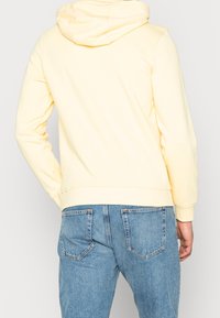 Person som bär en ljusgul hoodie och ljusblå jeans i denim sett bakifrån mot en enfärgad bakgrund.