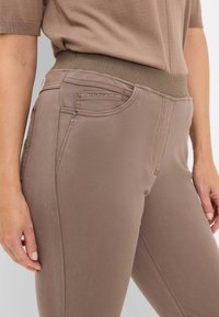 Slim-fit bukser i taupe stof med elastisk talje. Har en frontlomme med en dekorativ lynlås og subtile syninger.