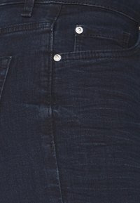 Los pantalones vaqueros de mezclilla azul oscuro presentan un diseño de cinco bolsillos con herrajes plateados, incluidos botón y remaches; textura suave con un ligero brillo.