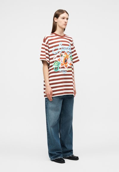 Γυναίκα που φοράει υπερμεγέθη καφέ και λευκή ριγέ μπλούζα με γραφική παράσταση φρούτων, χαλαρά μπλε τζιν και μαύρα παπούτσια, στέκεται μπροστά σε μια απλή φόντο.