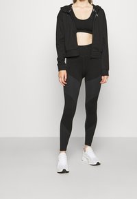 Zwarte sportoutfit bestaande uit een cropped hoodie en high-waisted leggings met gestructureerde accenten, gecombineerd met witte sportschoenen.