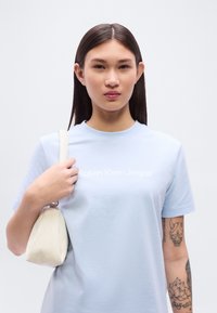 Ανοιχτό μπλε βαμβακερό t-shirt με το λογότυπο «Calvin Klein Jeans» σε λευκό. Το μοντέλο κρατά μια τσάντα ώμου κρεμ χρώματος, παρουσιάζοντας ένα καθαρά casual look.