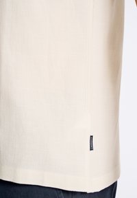Camisa de manga corta color crema con una tela suave y texturizada. Presenta una etiqueta en la costura lateral y un dobladillo recto. Diseño minimalista.