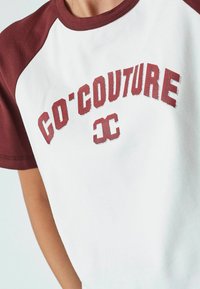 T-shirt bianco in cotone con maniche raglan color borgogna. Presenta la scritta "CO'COUTURE" stampata in lettere borgogna sbiadite sul petto.