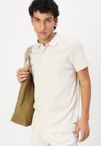 Polo shirt beige clair avec un motif texturé, à manches courtes et col. Tenant un sac fourre-tout marron, positionné contre un fond blanc.