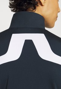 Giacca sportiva nera con colletto alto e un logo geometrico bianco prominente sul retro. Tessuto liscio e elasticizzato.
