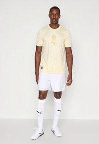 Šviesiai geltona sportinė marškinėliai su apvalia iškirpte ir logotipu, derinami su baltais šortais, baltais kojinėmis iki kelių ir baltais bateliais.