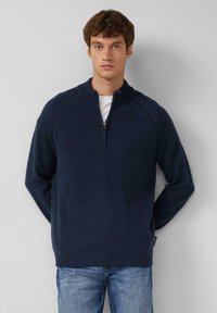 Granatowy sweter z zamkiem i ściągaczami na rękawach oraz u dołu, wykonany z miękkiej, strukturalnej tkaniny. Nosi się go na białej koszuli z dopasowanymi dżinsami.