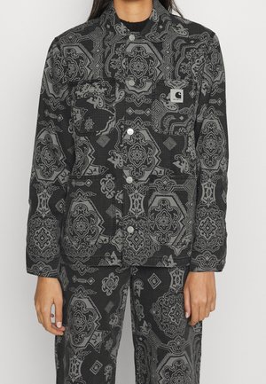 Musta takki ja housut, joissa harmaat monimutkaiset paisley-kuvioiset geometriset kuviot, metalliset painonapit ja Carharttin logopatch rinnataskussa.