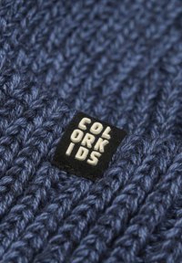 Marinblått stickat tyg med tjock textur; pryds av en fyrkantig svart etikett med texten "COLOR KIDS" i vitt, mot den stickade bakgrunden.