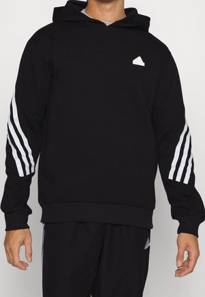 Mann trägt schwarzen Hoodie mit weißen Streifen an den Ärmeln und kleinem weißem Sneaker-Logo auf der Brust, kombiniert mit schwarzer Hose.