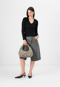 Eine Frau steht vor einem schlichten Hintergrund, trägt ein schwarzes Langarmshirt, graue Shorts und hält eine tasche mit Schlangen-Muster.