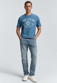 Sininen pyöreäkauluksinen t-paita valkoisella tekstillä, yhdistetty vaaleansinisiin farkkuihin ja valkoisiin tennareihin. Malli seisoo kädet taskuissa neutraalia taustaa vasten.