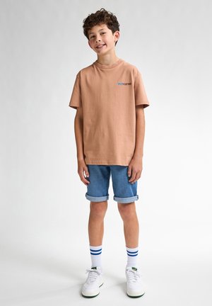T-shirt beige en coton à manches courtes avec un logo bleu. Associé à un short en denim bleu, des chaussures blanches et des chaussettes blanches avec des rayures bleues.