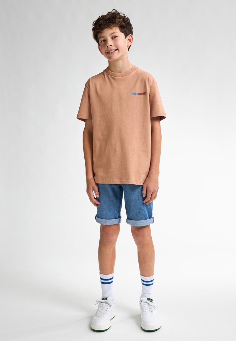 Camiseta de algodón beige con mangas cortas y logo azul. Combinada con pantalones cortos de mezclilla azul, zapatos blancos y calcetines blancos con rayas azules.