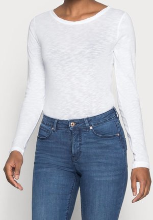 Frau trägt ein eng anliegendes weißes Langarmshirt, in hoch taillierte blaue Jeans gesteckt, dargestellt vom Hals bis zur Hüfte.