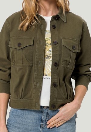 Kvinde iført olivengrøn knap-op jakke med klaplommer over hvid grafisk t-shirt og blå jeans, hånd hviler nær lommen.