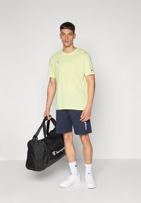 Lys grønn t-skjorte med "Champion" logo, marineblå shorts, hvite joggesko og svart treningsbag. Enkelt design, bomullsstoff, uformell stil.