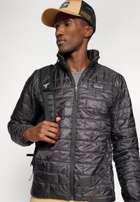 Veste matelassée noire avec fermeture éclair, col haut et poches latérales. Portée par-dessus une chemise grise, associée à une casquette beige et un sac à dos noir.