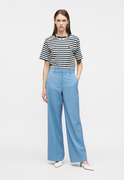 Γυναίκα όρθια με μαύρο και άσπρο ριγέ μπλουζάκι, ανοιχτό μπλε παντελόνι με φαρδιά ποδόγυρα, άσπρες ψηλοτάκουνες γόβες και κολιέ με μαργαριτάρια, μπροστά από απλό φόντο.