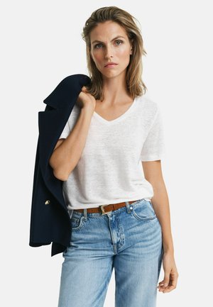 Donna con capelli biondi fino alle spalle indossa una T-shirt bianca a scollo a V e jeans blu, tenendo una giacca blu navy sulla spalla su sfondo bianco.