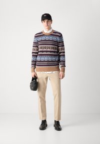 Strickpullover mit einem mehrfarbigen Fair-Isle-Muster, beigebrauner Rippbund, kombiniert mit beigen Hosen und schwarzen Schuhen; hält eine schwarze Tasche.
