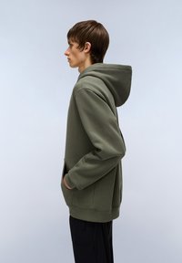 Sweatshirt olive vert avec une poche kangourou, tissu texturé et capuche avec cordon de serrage réglable, porté avec un pantalon noir à jambe droite.