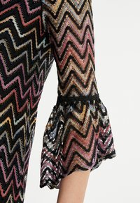 Robe noire transparente présentant un motif en chevrons aux couleurs vives, avec des poignets en dentelle évasés et un design texturé.