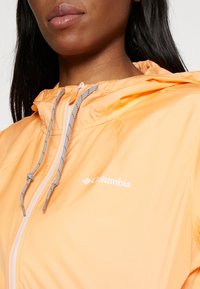 Light orange, lightweight jacket with a hood, featuring gray drawstrings and a zipper, displaying the Columbia logo on the left chest.