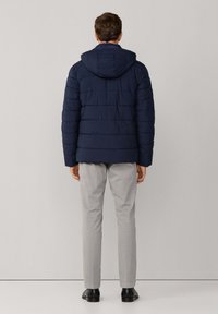 Chaqueta acolchada azul marino con capucha, con un patrón acolchado horizontal, combinada con pantalones gris claro y zapatos negros.