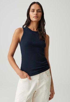 Femme aux longs cheveux noirs portant un haut sans manches bleu marine et un pantalon blanc, debout avec une main dans la poche devant un fond uni.