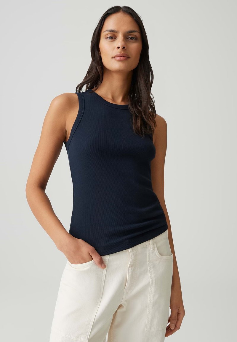 Vrouw met lang donker haar draagt een marineblauwe top zonder mouwen en witte broek, staand met één hand in haar zak tegen een effen achtergrond.