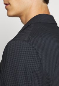 Blazer scuro con texture e un sottile motivo a fantasma. Il colletto è di stile classico a rever, che mette in risalto una vestibilità su misura e un tessuto liscio.
