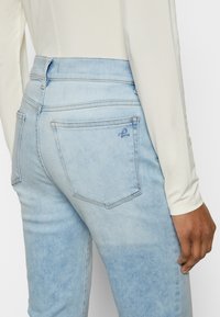 Jean en denim bleu clair à taille haute, présentant un logo brodé discret sur la poche arrière et des détails de couture standard.