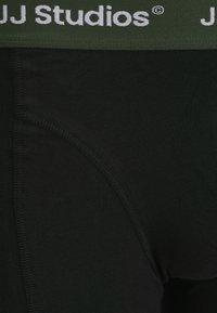 Schwarze Stoff-Boxershorts mit einem grünen elastischen Bund und dem "JJ Studios"-Logo. Glatte Textur und genähte Nähte für Langlebigkeit.