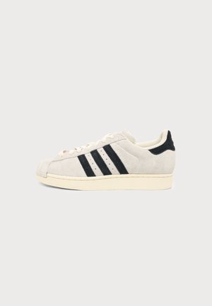 Baskets basses en daim beige avec des bandes noires sur le côté et une semelle en caoutchouc crème sur fond clair uni.