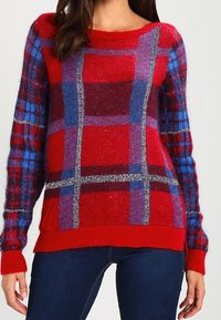 Frau trägt einen gestrickten Pullover mit rot-blau-lila Karomuster, langen Ärmeln und dunkelblauen Jeans.