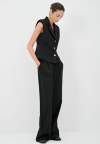 Schwarzer ärmelloser Blazer mit einer figurbetonten Passform, goldenen Knöpfen, kombiniert mit lockeren schwarzen Hosen, was einen eleganten, maßgeschneiderten Look schafft.