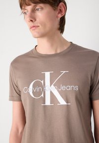 Ruda medvilnės marškinėliai su trumpomis rankovėmis, pasipuošę dideliu baltu logotipu "Calvin Klein Jeans" krūtinės srityje, apvaliu kaklu, glotnia tekstūra.