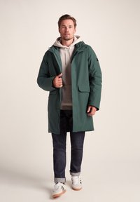 Green waterproof coat with a hood, zippered front, and two front pockets. Worn over a light hoodie, paired with dark jeans and sneakers.