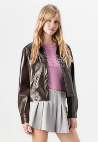 Brun kunstlæder cropped jakke med knaplukning, brystlomme og lange ærmer, sammen med en pink top og en grå plisseret nederdel.