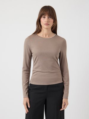 Donna con capelli castani lisci che indossa un top lungo aderente color taupe e pantaloni neri a gamba larga, in piedi contro uno sfondo neutro.