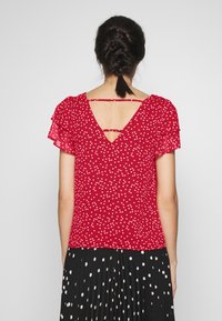 Rød T-shirt med korte ærmer og hvide hjerte mønstre, flæseærmer og V-rygdesign med en lille udskæring og stropdetaljer.