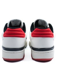 Hvite sneakers med svart kanting, røde hælaccenter og teksturerte såler. Har en polstret krage og tydelig logo bak.