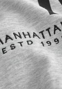 Tela gris con una textura suave, con texto bordado en negro que dice "MANHATTAN ESTD 1993". Una toma en primer plano resalta los detalles del bordado.
